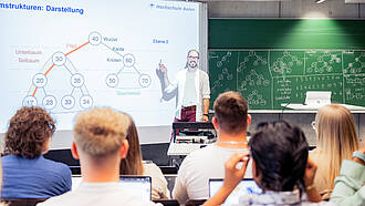 Ein gut besetzter Vorlesungssaal. Man sieht die Studierenden von hinten. Vor ihnen erklärt ein Lehrender Stoff an einem Whiteboard. Auf dem Whiteboard und an der Tafel im Hintergrund sind Baumdiagramme zu sehen. Ein gut besetzter Vorlesungssaal. Man sieht die Studierenden von hinten. Vor ihnen erklärt ein Lehrender Stoff an einem Whiteboard. Auf dem Whiteboard und an der Tafel im Hintergrund sind Baumdiagramme zu sehen.