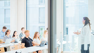 Man blickt seitlich in einen hellen Vorlesungsraum. Eine junge Professorin erklärt einigen Studierenden einen Sachverhalt. Im Hintergrund sieht man große Fenster, durch die ein weiteres Gebäude zu sehen ist. Man blickt seitlich in einen hellen Vorlesungsraum. Eine junge Professorin erklärt einigen Studierenden einen Sachverhalt. Im Hintergrund sieht man große Fenster, durch die ein weiteres Gebäude zu sehen ist.