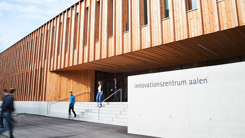 Auf dem Bild sieht man ein Gebäude aus Beton und Holz, über dessen Eingangstreppe Menschen das Gebäude verlassen. Auf dem Bild sieht man ein Gebäude aus Beton und Holz, über dessen Eingangstreppe Menschen das Gebäude verlassen.
