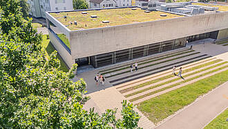 Das Aula- Hörsaalgebäude von oben. Auf dem Vorplatz unterhalten sich Studierende. Das Aula- Hörsaalgebäude von oben. Auf dem Vorplatz unterhalten sich Studierende.