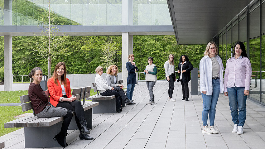 Vor einem Gebäude mit Glassteg in ein anderes Gebäude sind mehrere Frauen zu sehen, die in kleinen Grüppchen in die Kamera schauen. Vor einem Gebäude mit Glassteg in ein anderes Gebäude sind mehrere Frauen zu sehen, die in kleinen Grüppchen in die Kamera schauen.